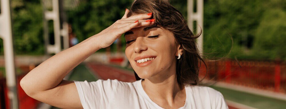 Mulher branca e de cabelos castanhos protegendo o rosto do sol para evitar o envelhecimento cutaneo Fotoprote&ccedil;&atilde;o